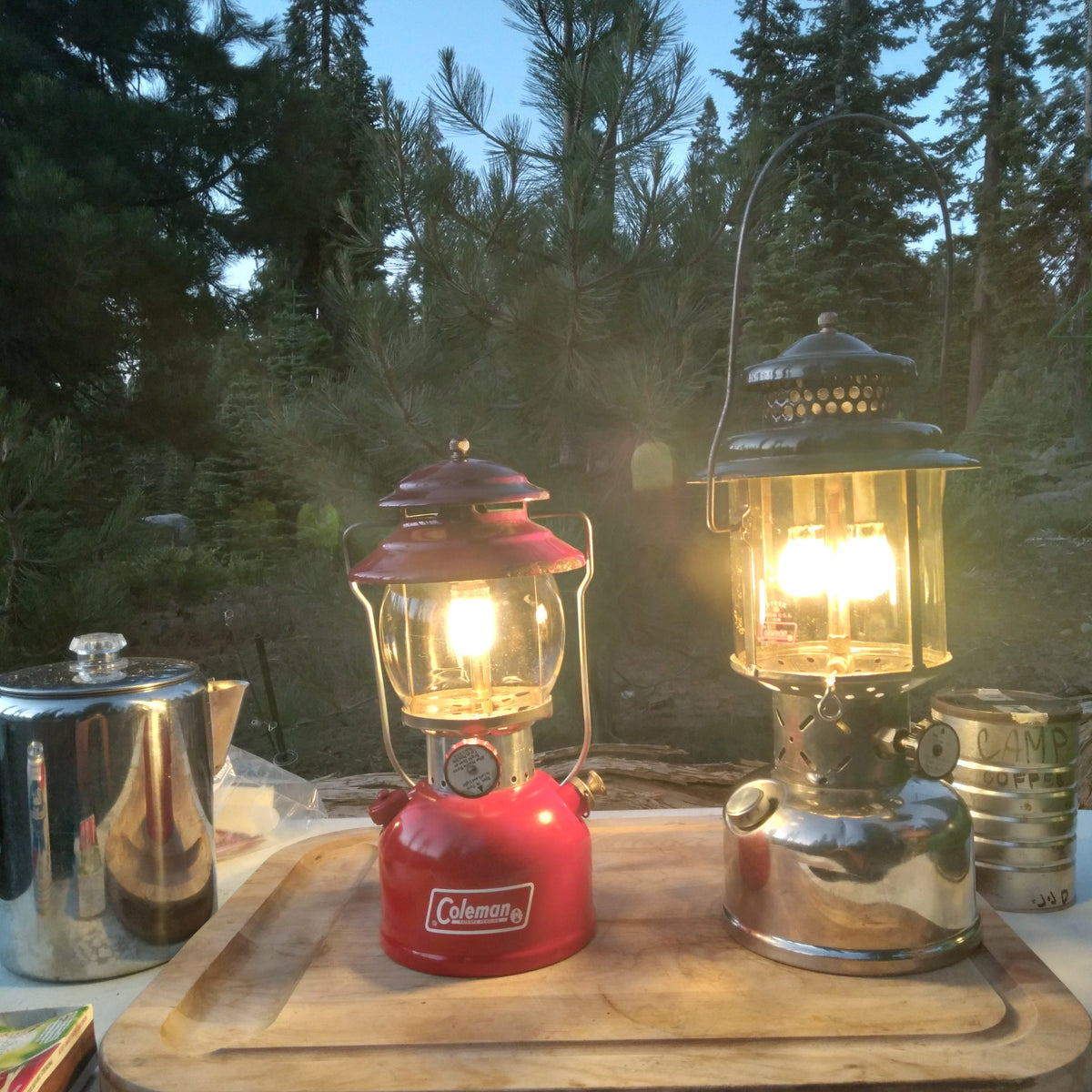 Two vintage Coleman lanterns, one red and one silver, lit and displayed on a wooden cutting board outdoors with trees in the background.